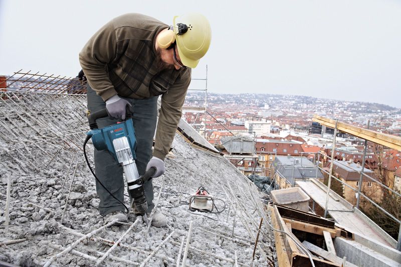A person wearing safety equipment uses a demolition hammer to break concrete on a rooftop.