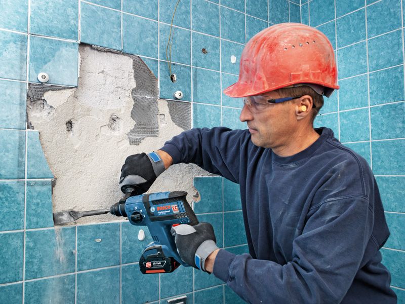 A person wearing safety equipment uses a cordless rotary hammer to remove wall tiles.