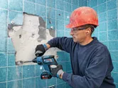 A person wearing safety equipment uses a cordless rotary hammer to chisel tiles from a wall.