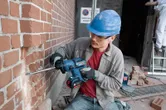 A person wearing safety equipment drills into a brick wall with a cordless rotary hammer.