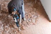 A person wearing safety equipment breaks floor tiles using a demolition hammer.