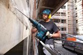 A person wearing safety equipment uses a demolition hammer to break concrete on a building site.