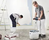 A person wearing safety equipment uses a stirring mechanism to mix paint in a bucket.