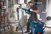 A person wearing safety equipment drills into a wall near pipes using a rotary hammer.