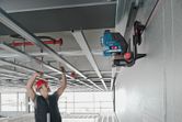 A person wearing safety equipment installs ceiling framing using a laser leveling tool on a wall mount.
