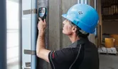 A person wearing safety equipment scans a concrete wall with a wallscanner detector.