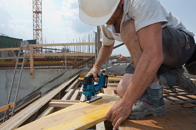 A person wearing safety equipment uses a jigsaw to cut wood at a construction site.