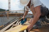A person wearing safety equipment uses a jigsaw to cut wood at a construction site.