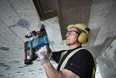 A person wearing safety equipment drills into a concrete ceiling with a cordless rotary hammer.