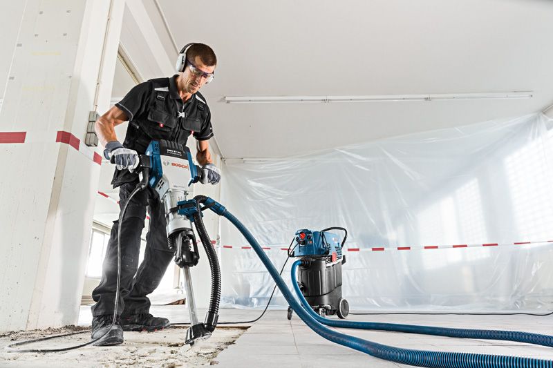A person wearing safety equipment uses a demolition hammer with dust extraction on a concrete floor.