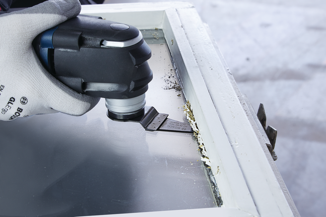 Worker wearing safety equipment removes old paint from a window frame using a multi-tool.