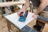 A person sands a wooden board with a cordless orbital sander on a workbench.