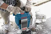 A person wearing safety equipment operates a demolition hammer to break concrete.