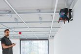 A person operates a laser leveling tool mounted on a wall to align ceiling framework.