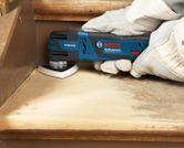A person wearing safety equipment sands a wooden stair with a cordless multi-cutter.