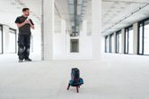 A person stands in a large empty room while a laser leveling tool accessory projects a green line.