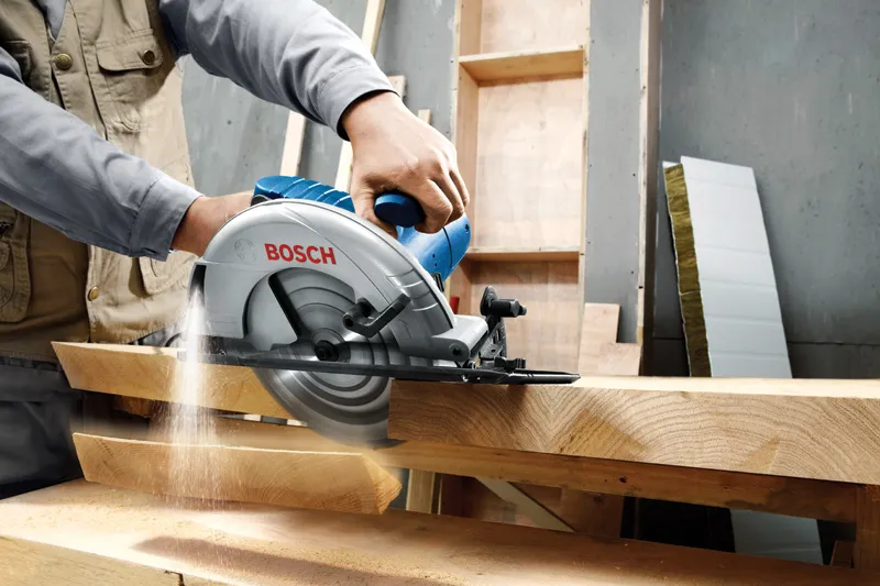 A person guides a circular saw through thick wooden boards in a workshop.