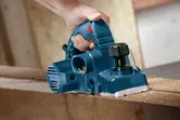A person uses a blue planer to smooth the surface of a wooden beam.