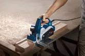 A person uses a planer to smooth wooden planks on a large workbench.