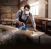 A person wearing safety equipment smooths a wooden beam with an electric planer.