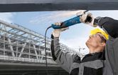 A person wearing safety equipment grinds overhead metal, producing sparks.