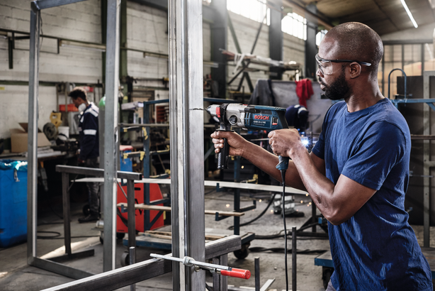 Person wearing safety equipment drills into a metal frame in an industrial workshop.
