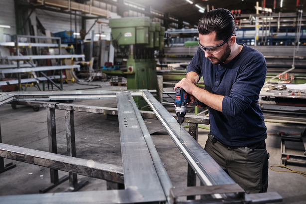 A worker wearing safety equipment drills holes into a metal frame in a factory.