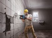 A person wearing safety equipment drills into a tiled wall in a partially demolished room.