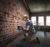 A person wearing safety equipment drills into a brick wall inside a building under construction.