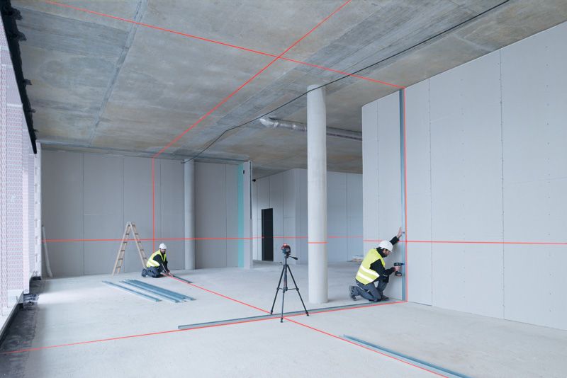 Two persons wearing safety equipment align drywall using a laser leveling tool in a building.