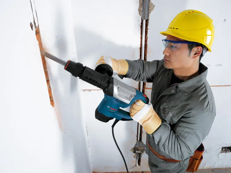 A person wearing safety equipment uses a demolition hammer to break a groove in a wall.