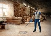 A person wearing safety equipment uses a blower to clear sawdust in a woodworking shop.