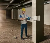 A person wearing safety equipment drills into a concrete pillar in a construction area.
