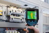 Person uses a thermo camera to inspect electrical panels for heat anomalies.