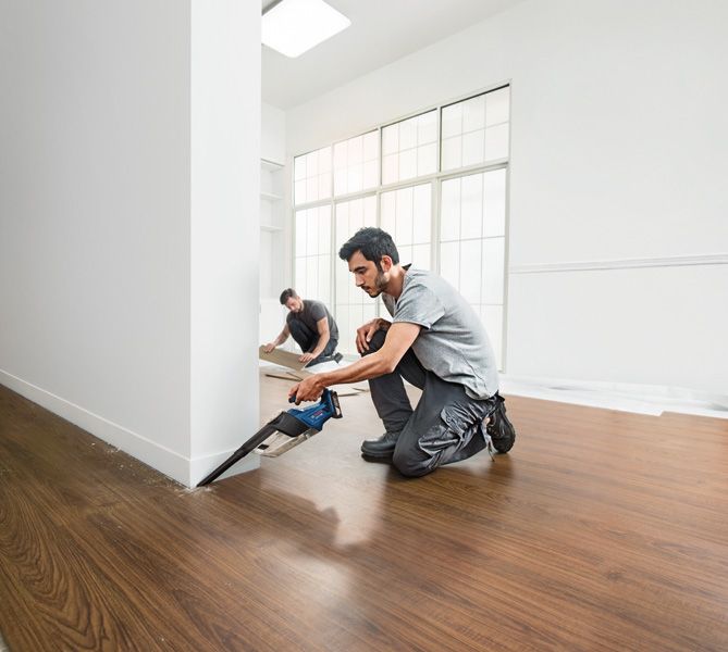 A person cuts flooring near a wall with a cordless saw while another installs floor panels.