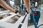 Person using a power router to shape a wooden panel in a workshop.