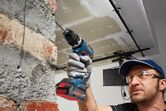 A person wearing safety equipment drills into a brick wall with a cordless power drill.