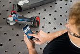 A person wearing safety equipment uses a smartphone to control a cordless angle grinder.