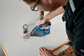 A person wearing safety equipment cleans debris near an electrical outlet with a cordless vacuum.