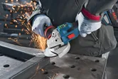 A person wearing safety equipment grinds a metal beam with a cordless angle grinder.