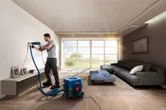 A person drills into a living room wall using a power tool connected to a dust extractor.