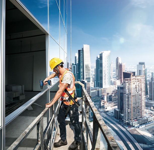A person wearing safety equipment uses a power drill on a high-rise building balcony.