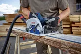Person wearing safety equipment cuts a rough wooden plank with a circular saw.