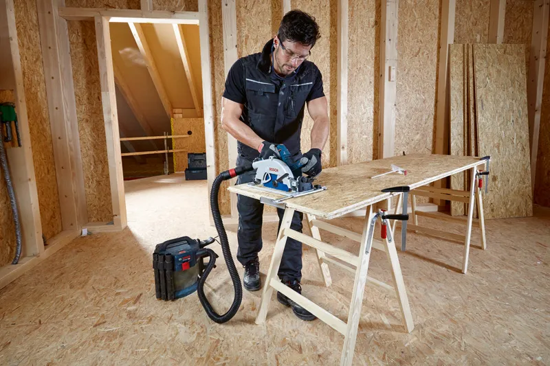 A person wearing safety equipment cuts plywood with a circular saw connected to a dust extractor.