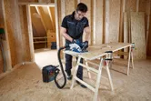 A person wearing safety equipment cuts plywood with a circular saw connected to a dust extractor.