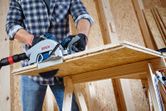 A person wearing safety equipment uses a circular saw to cut a wooden board.