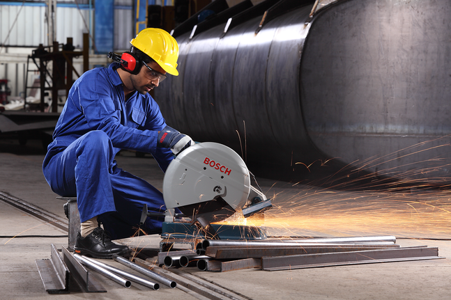 Person wearing safety equipment cuts metal pipes with a power saw in an industrial setting.