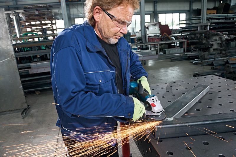 GWS 18V-10 A person wearing safety equipment grinds a metal beam with an angle grinder in a workshop.