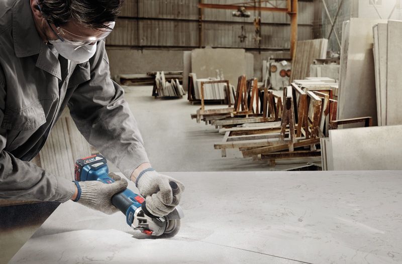 GWS 18V-10 A person wearing safety equipment cuts a slab with a cordless angle grinder in a workshop.