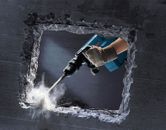 A person wearing safety equipment uses a demolition hammer to break a concrete wall.